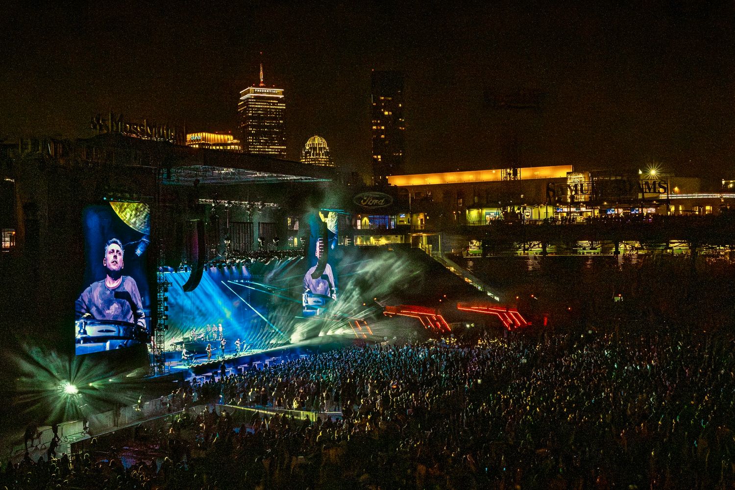 A large outdoor concert crowd illuminated by vibrant blue stage lights and video screens in a nighttime arena setting.
