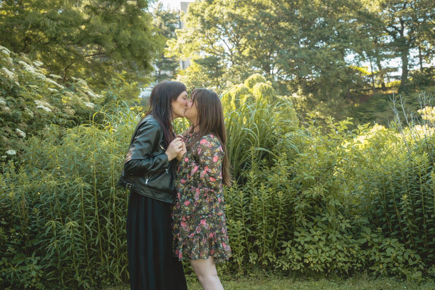 Two women sharing a romantic kiss outdoors surrounded by lush green foliage and trees.