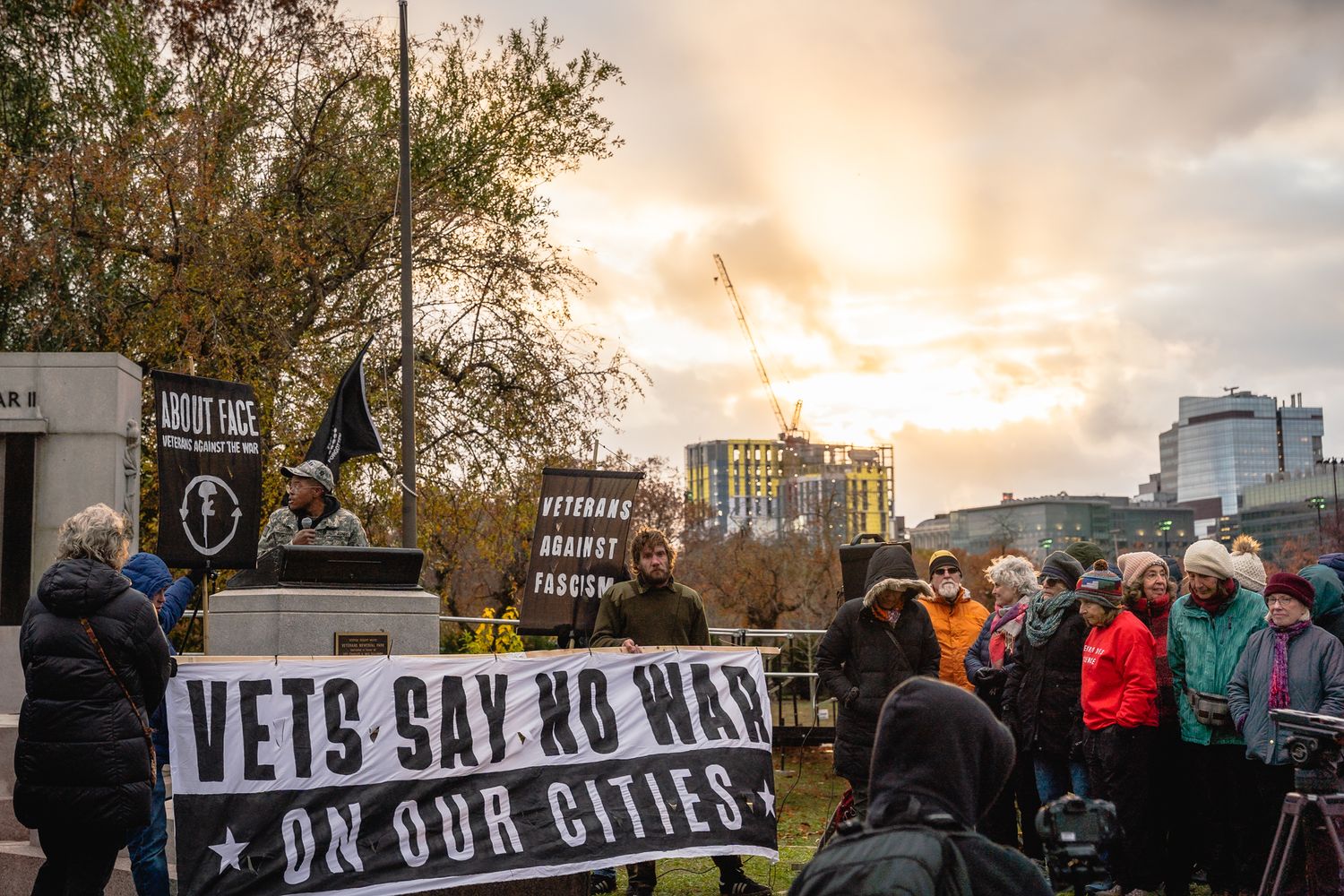 A panoramic view of protesters holding signs at a rally during sunset with buildings visible in the background.