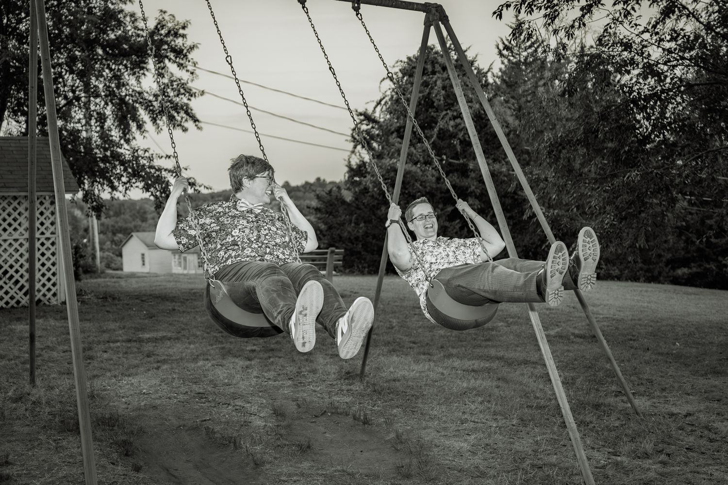A vintage black and white photograph shows two people enjoying a swing set outdoors in a backyard setting.
