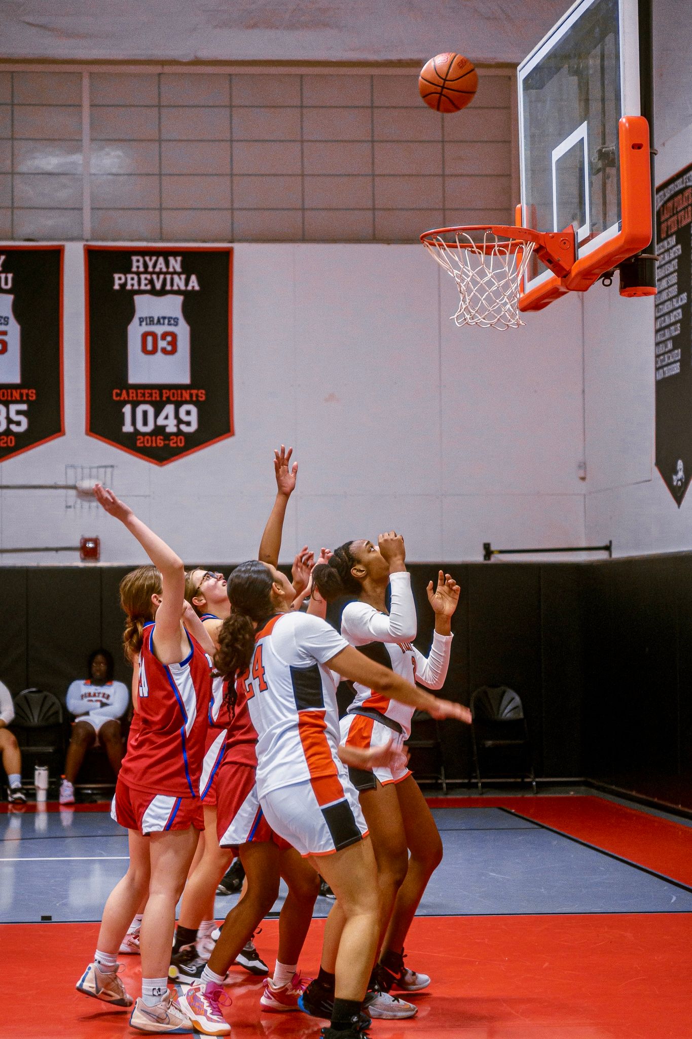 Basketball players compete for a rebound under the hoop on a red court during an indoor game.