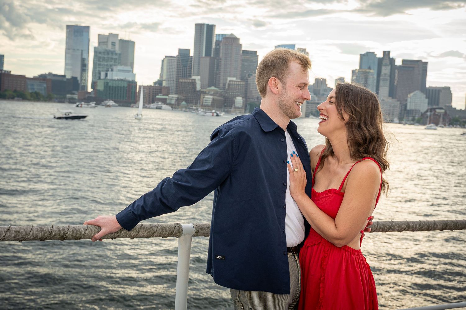 Couple embracing and smiling by the waterfront with Boston city skyline in the background on a cloudy day.