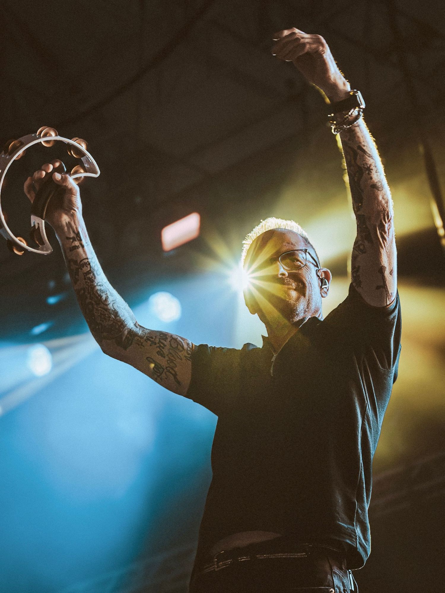 Tattooed performer raises arms triumphantly on stage holding tambourine with dramatic blue and yellow stage lighting behind him.