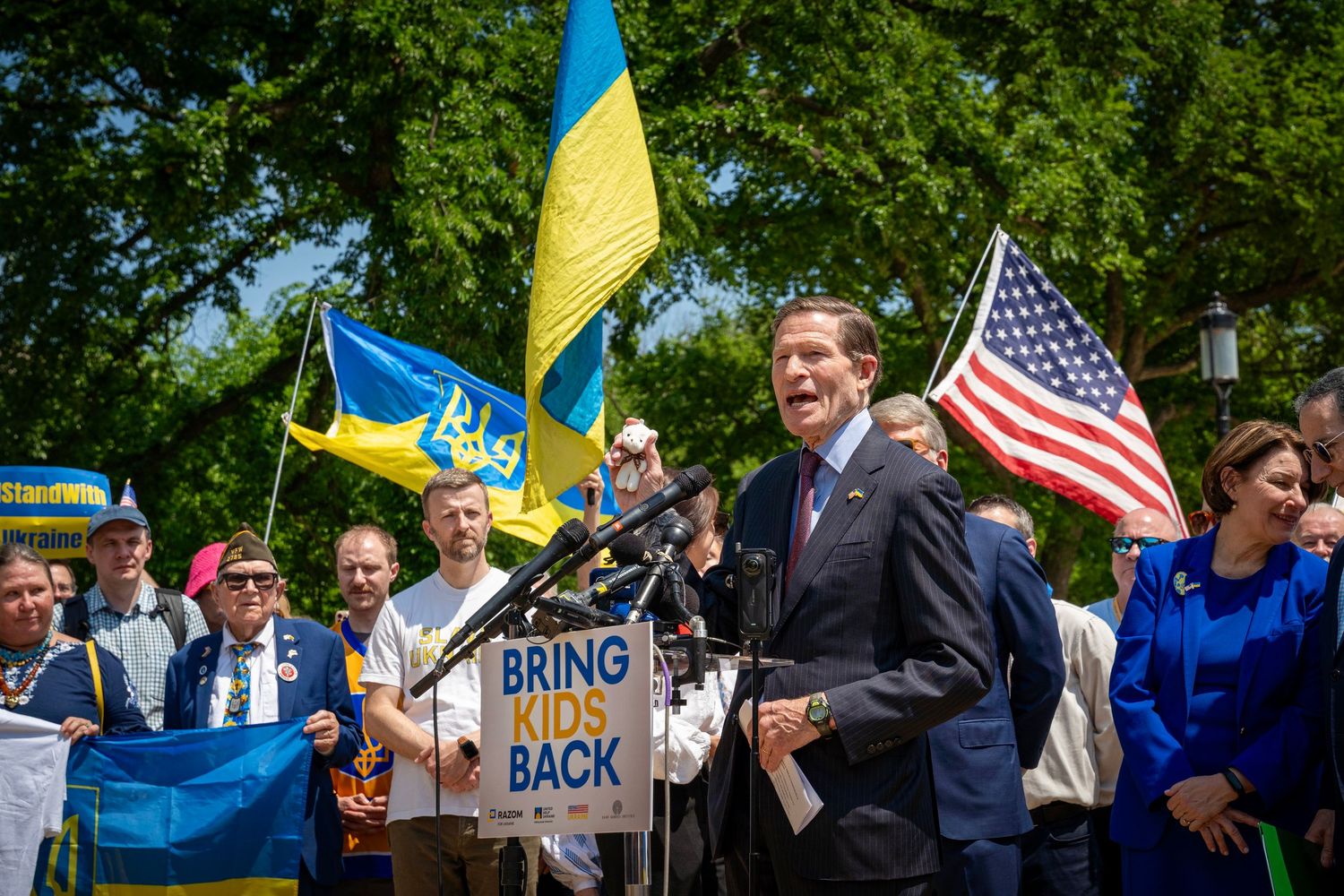 Politician speaks at outdoor rally with Ukrainian and American flags, crowd holds 'Bring Kids Back' sign.