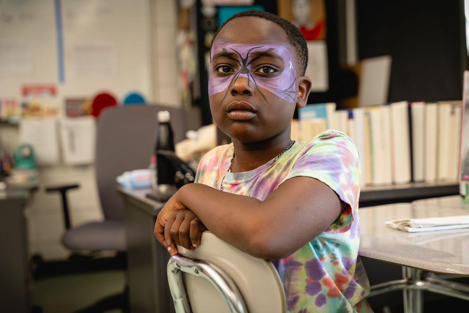 Child with purple face paint sits arms crossed in a colorful tie-dye shirt inside a classroom.