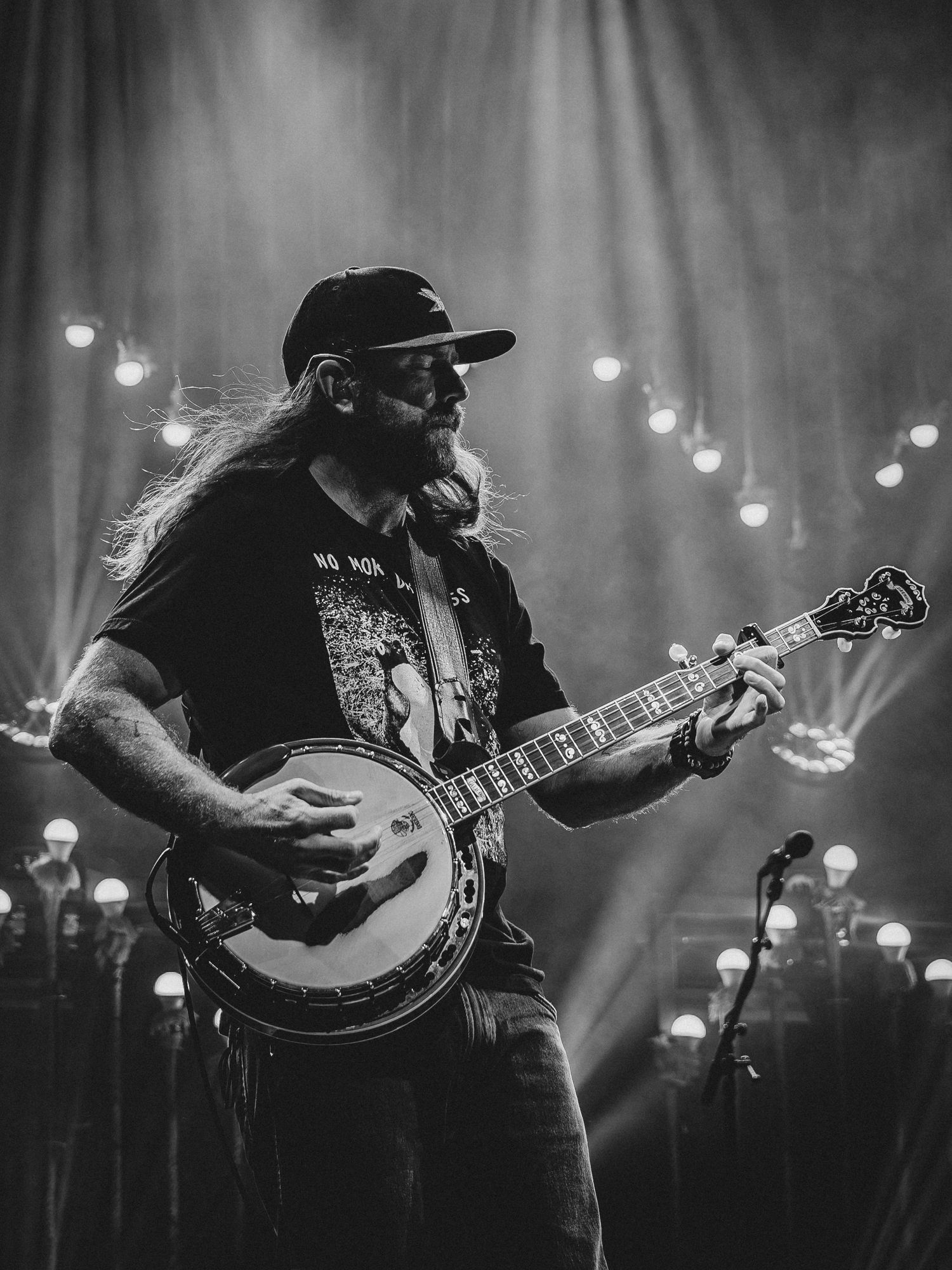 Bearded musician playing banjo on stage in black and white, wearing cap and graphic tee under dramatic concert lighting.