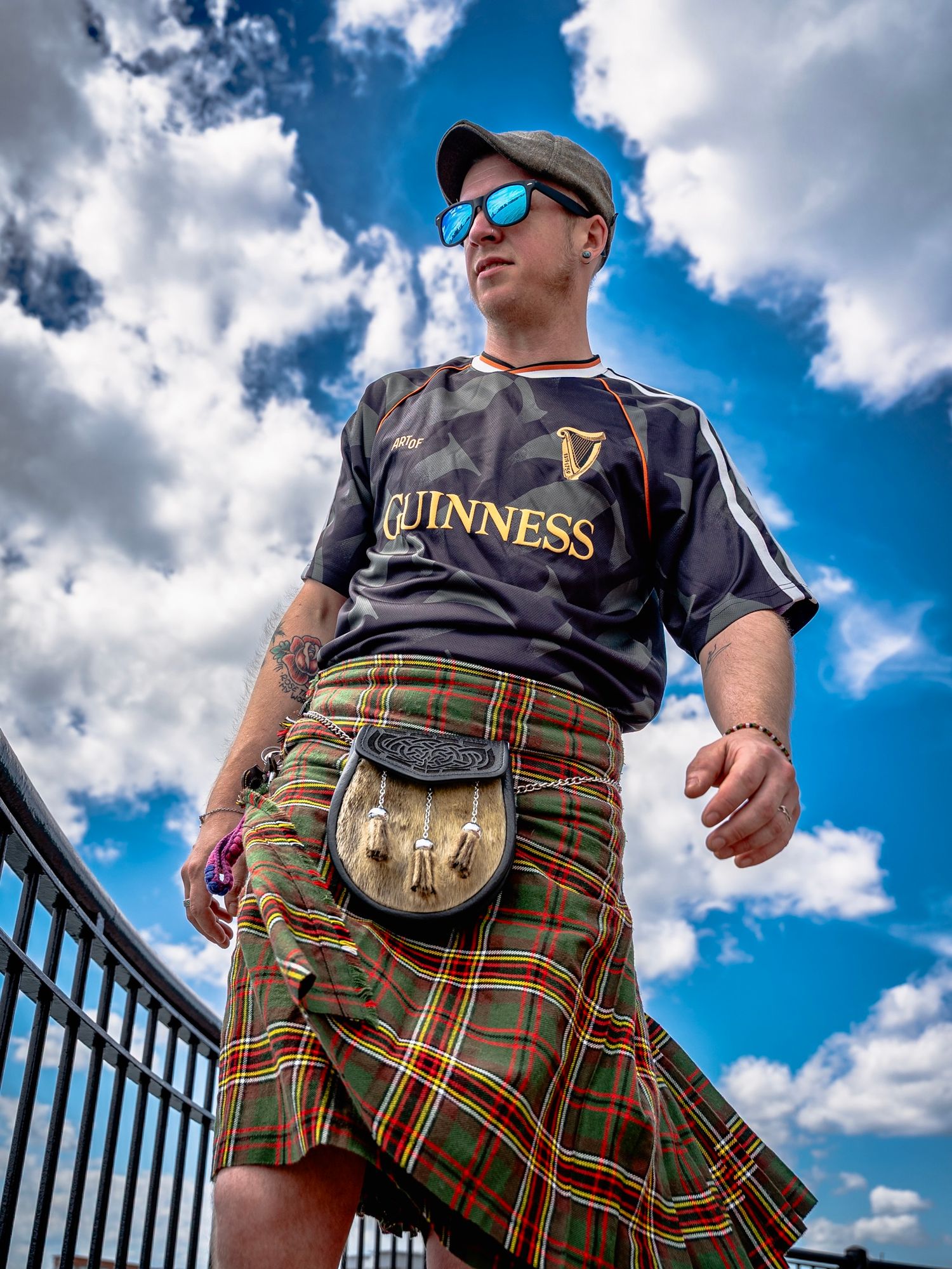 Man in Guinness jersey, plaid kilt, sporran, cap, and mirrored sunglasses poses confidently under a blue cloudy sky.
