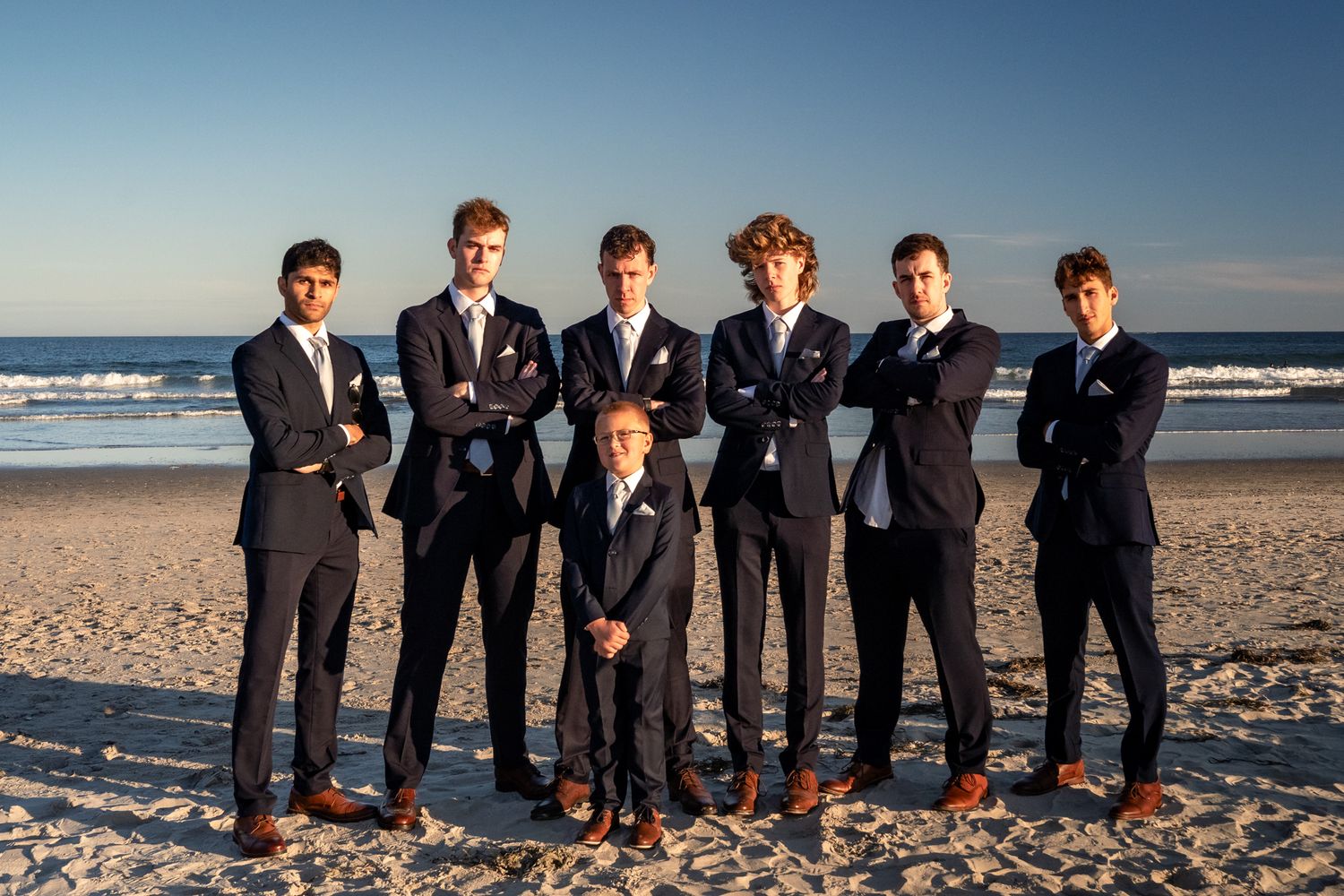 A wedding party in formal black suits poses together on a sunny beach during golden hour.
