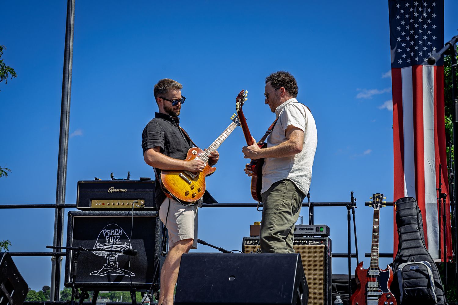 Two musicians perform on an outdoor stage with guitars and amplifiers against a blue sky and American flag backdrop.