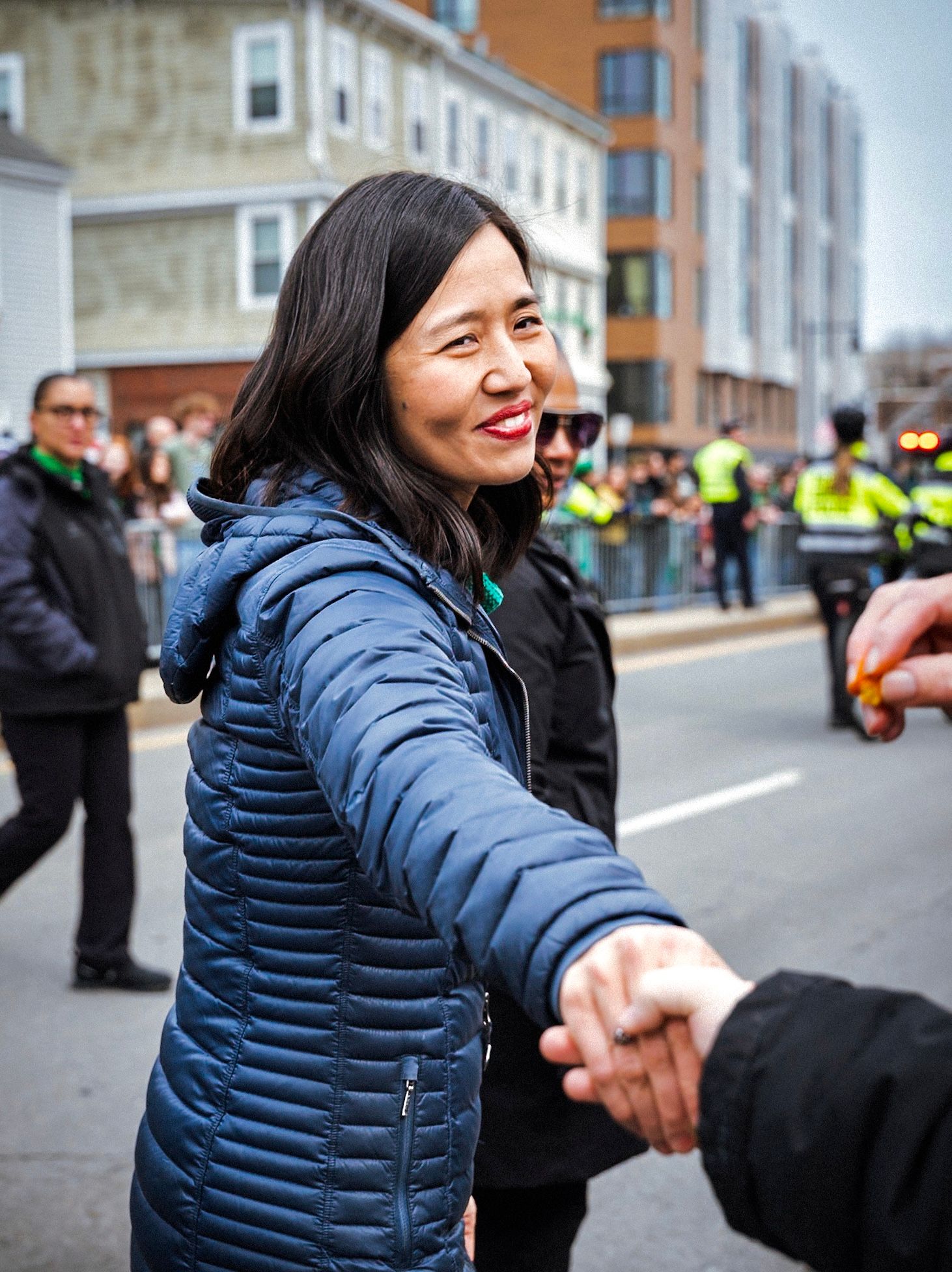 A smiling woman in a blue puffer jacket shakes hands at a festive street parade with crowds in the background.