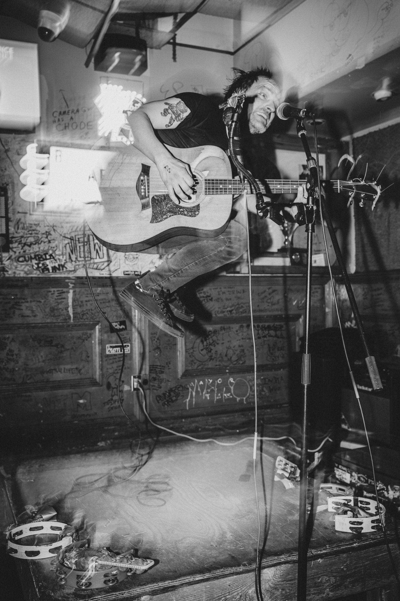 Black and white photo of a punk guitarist jumping mid-air on stage while playing acoustic guitar at a live music venue.