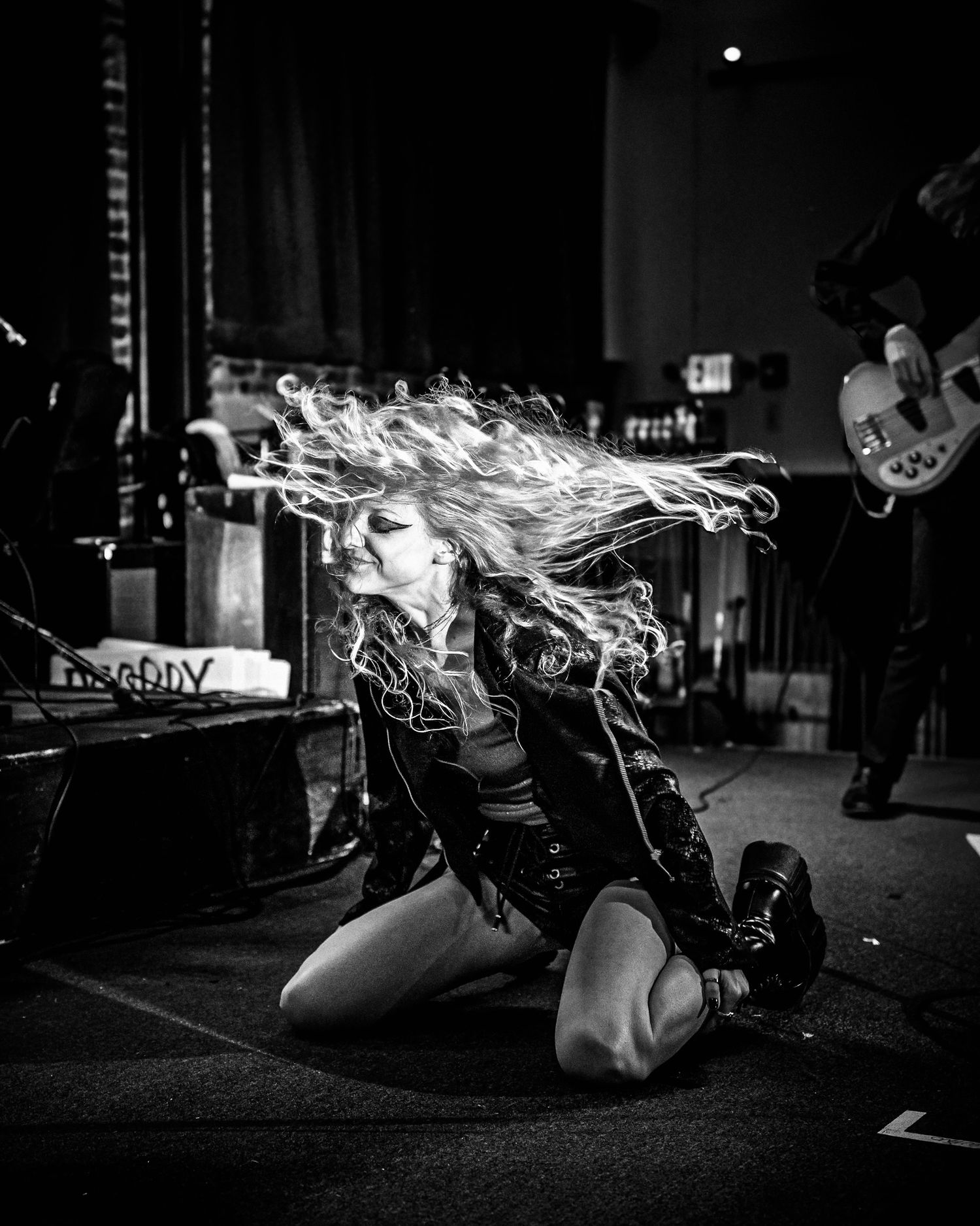 A performer in a dark concert venue whips their flowing hair while kneeling dramatically on stage during a music performance.