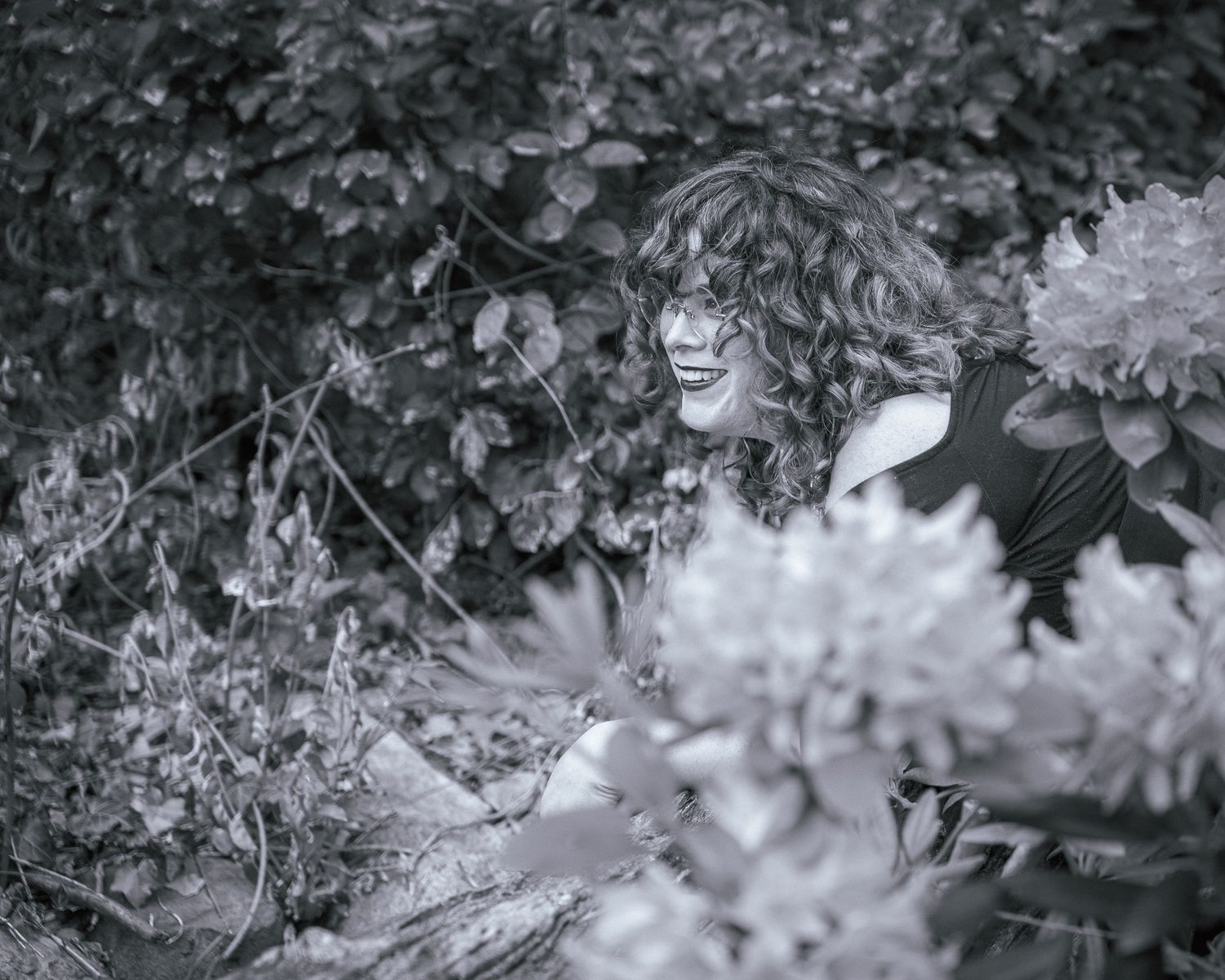 A black and white portrait photograph showing a joyful expression through foliage and flowers in a garden setting.