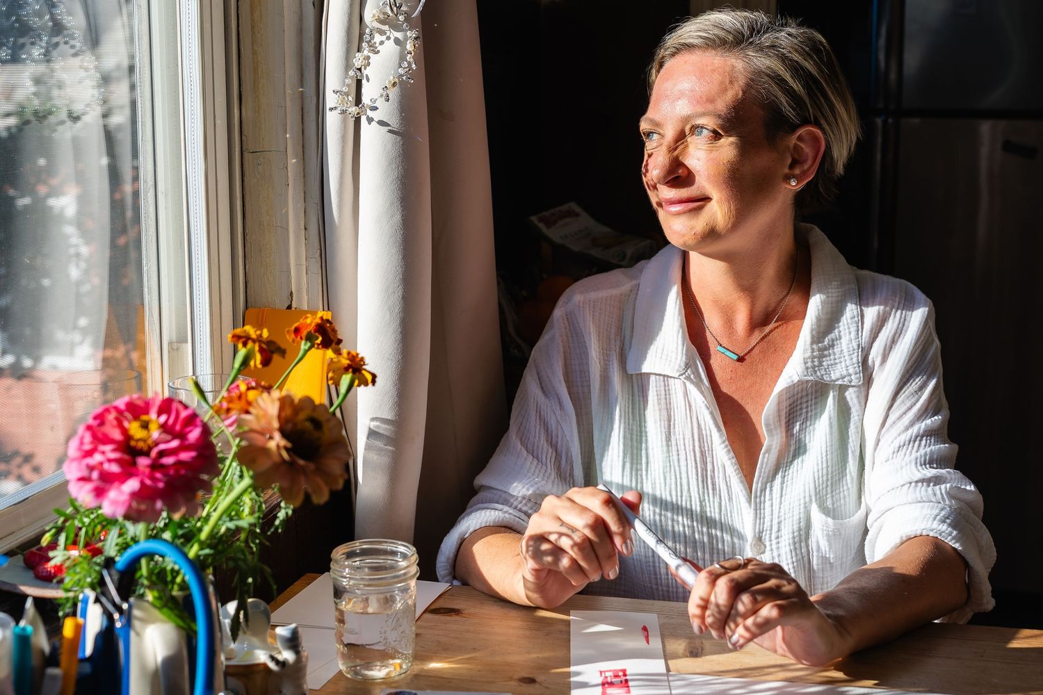 Woman painter sitting at sunny table with flowers, writing notes, wearing white shirt, looking thoughtfully out window.