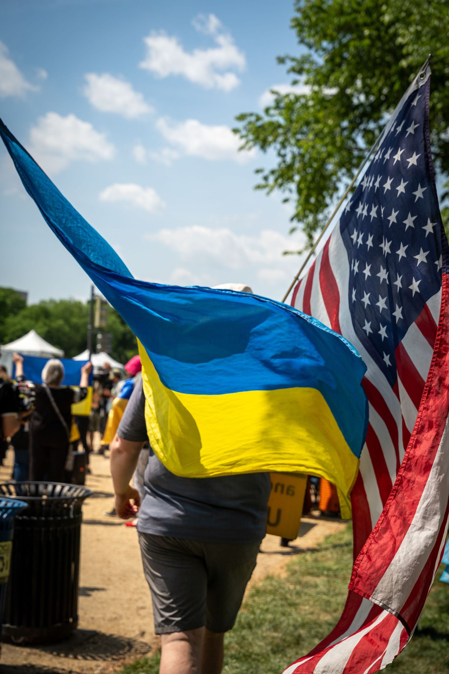 Person carrying Ukrainian and American flags at an outdoor rally on a sunny day.