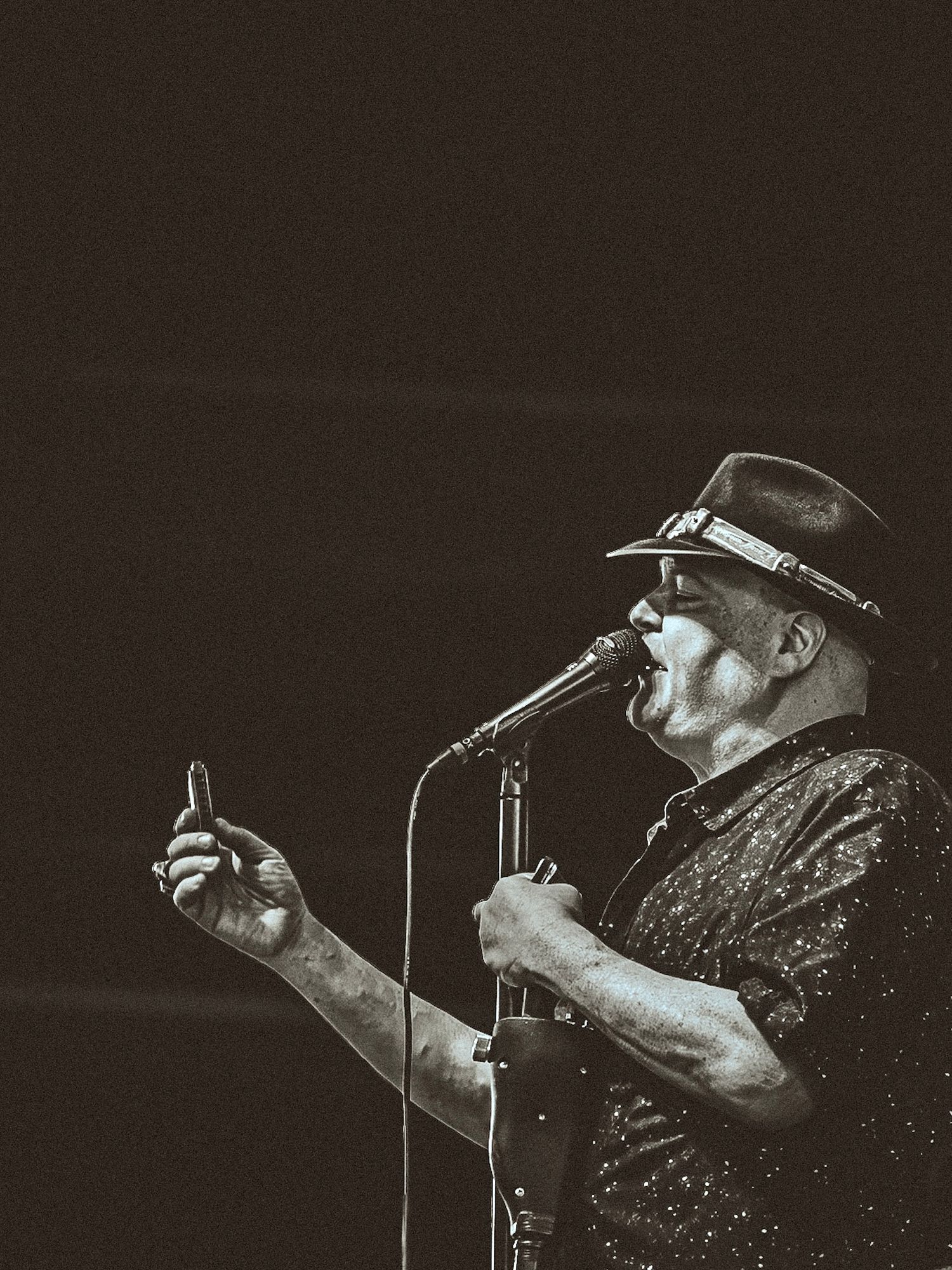Older male singer performing on stage wearing a hat and sunglasses, singing into a microphone in black and white.