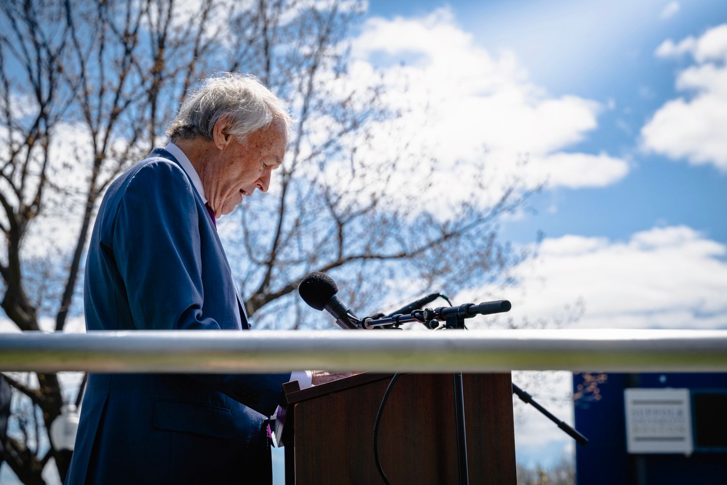 Elderly person speaking at outdoor podium under blue sky with clouds, wearing blue jacket at public event.