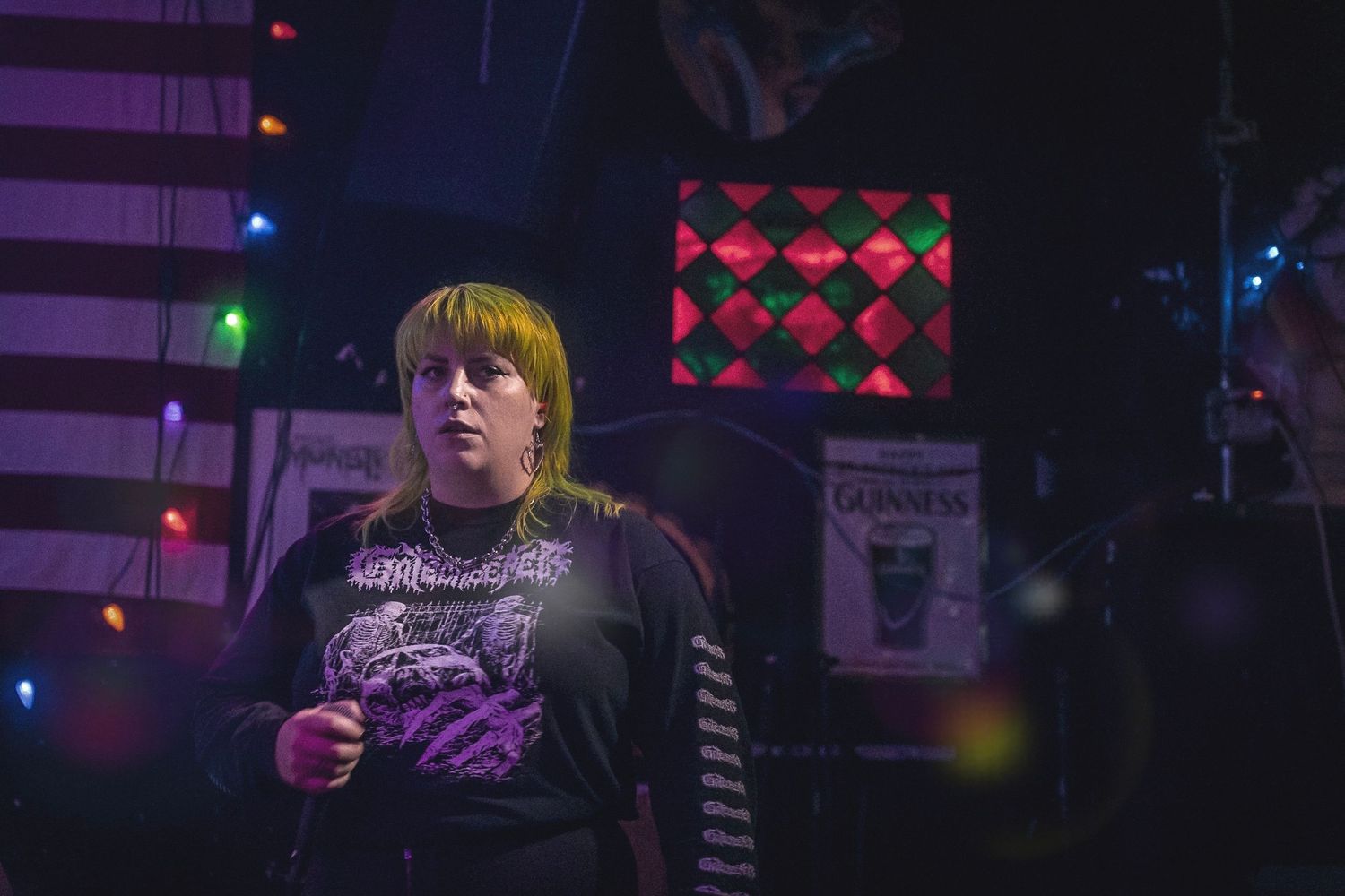 Blonde performer in graphic tee on dimly lit stage with colorful lights and Guinness sign in background.