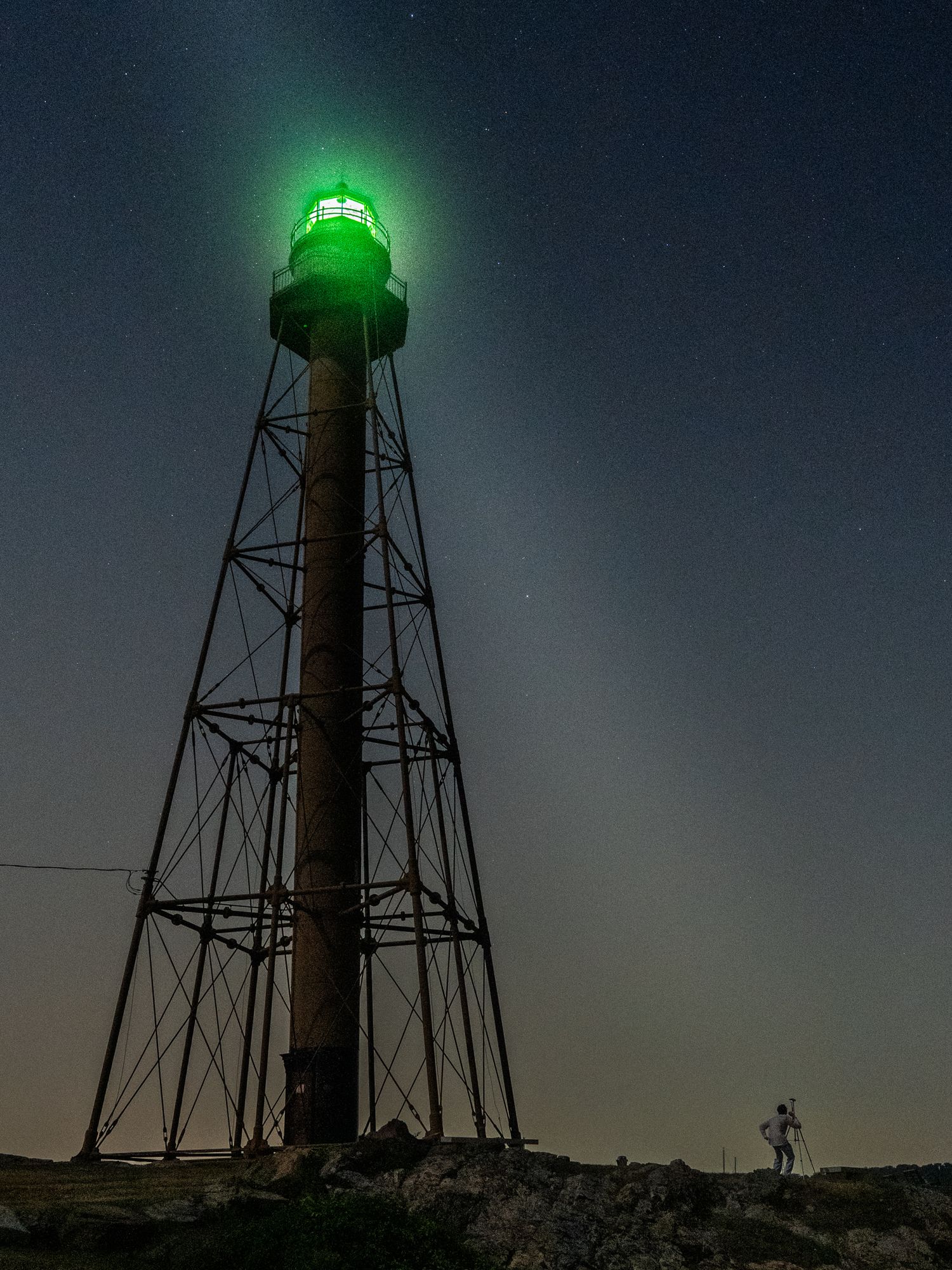 A tall lighthouse with a glowing green beacon illuminates the dark night sky while a small figure stands silently below.