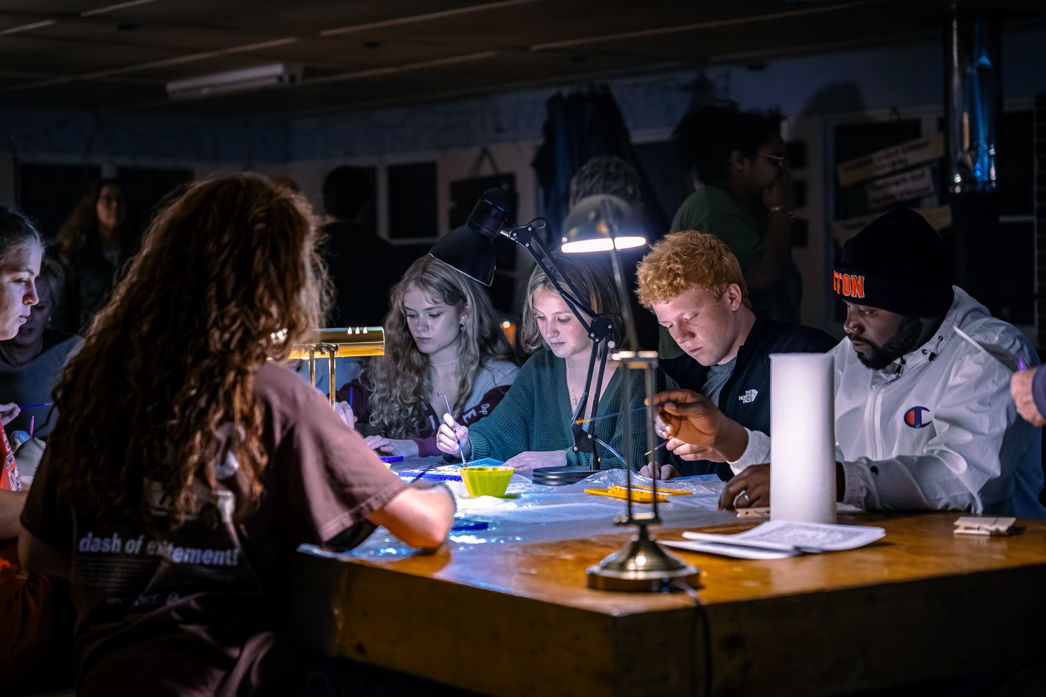 Students study together at a dimly lit table using phones and laptops in the evening.