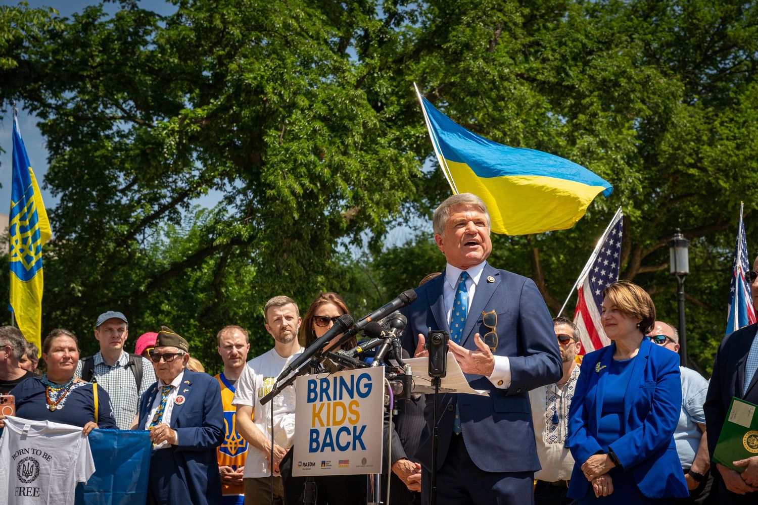 Politician speaks at outdoor rally with Ukrainian flags and 'Bring Kids Back' sign, crowd gathered behind.
