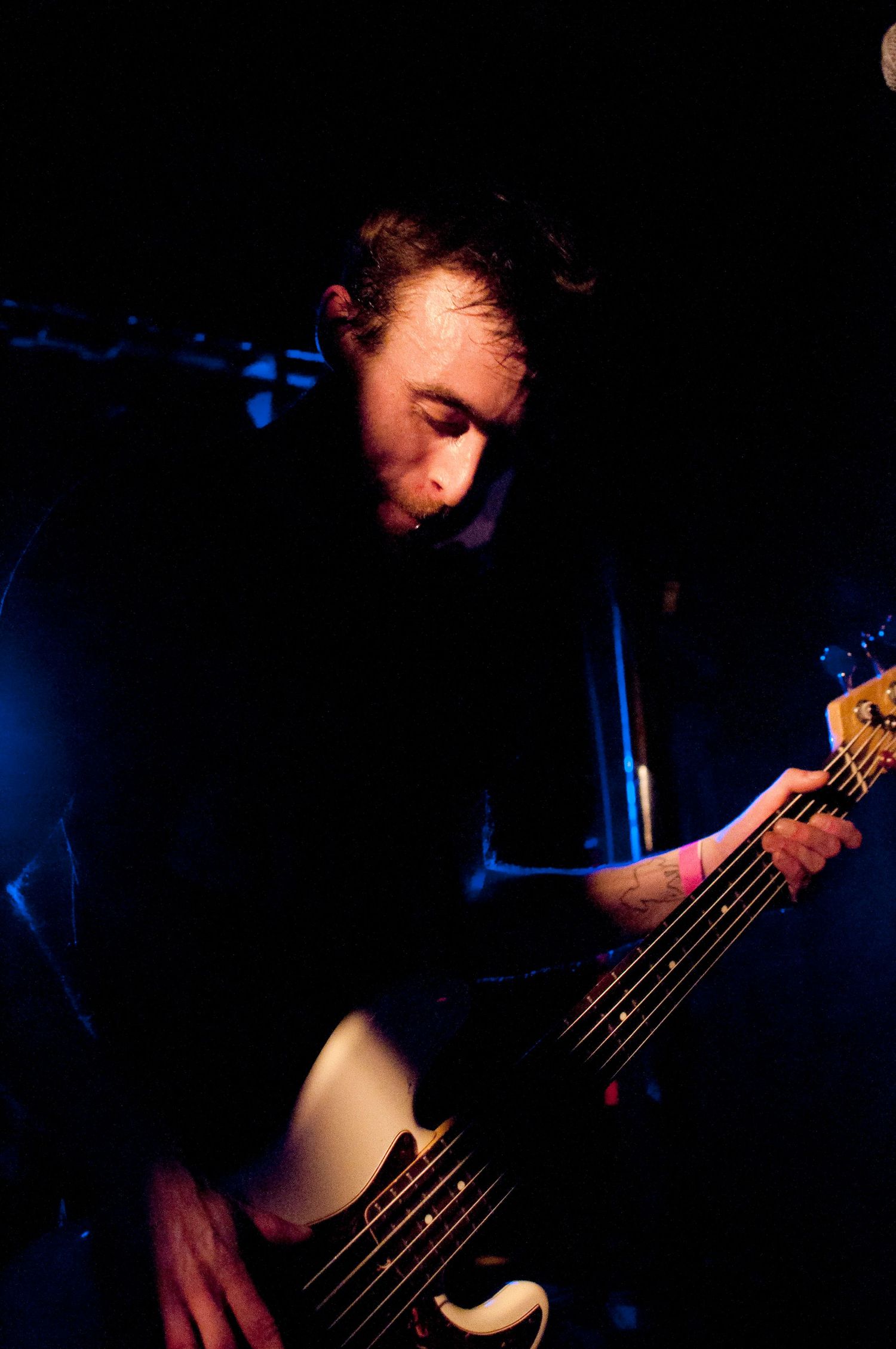 Bass guitarist plays in dramatic blue and red stage lighting while looking down at instrument during performance.