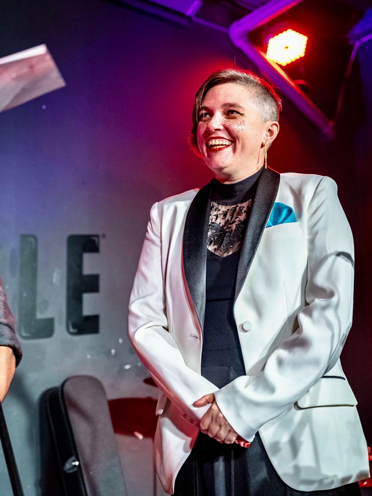 A performer in a white blazer and black shirt smiles while entertaining on stage under purple and red lighting.