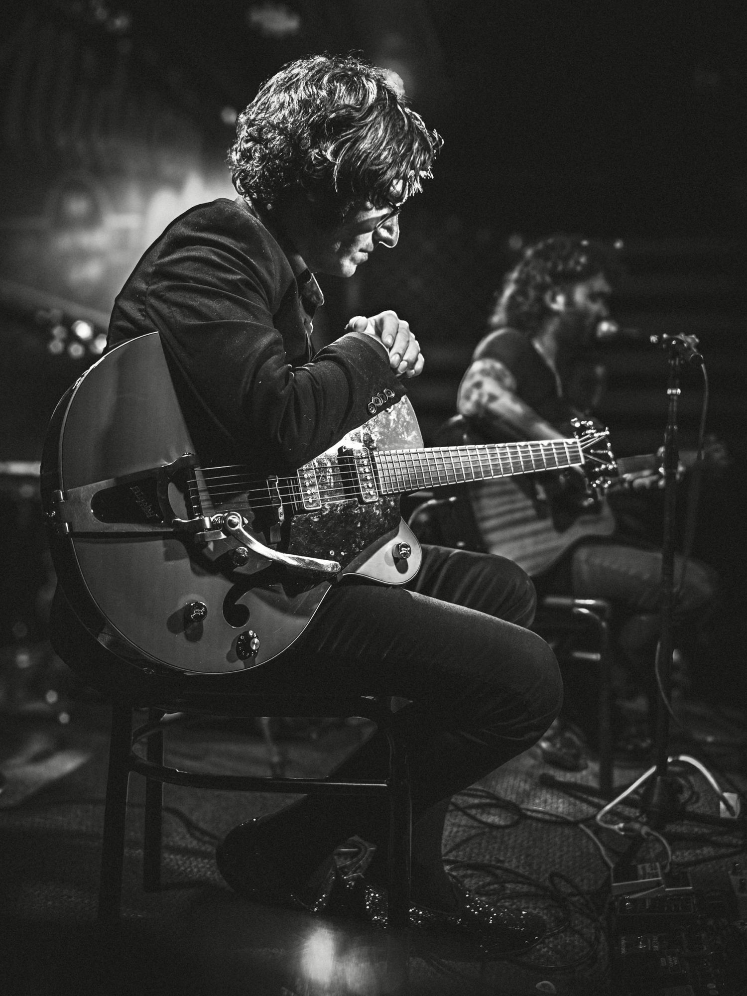 Black and white photo of a guitarist sitting and playing an electric guitar on stage during a live performance.