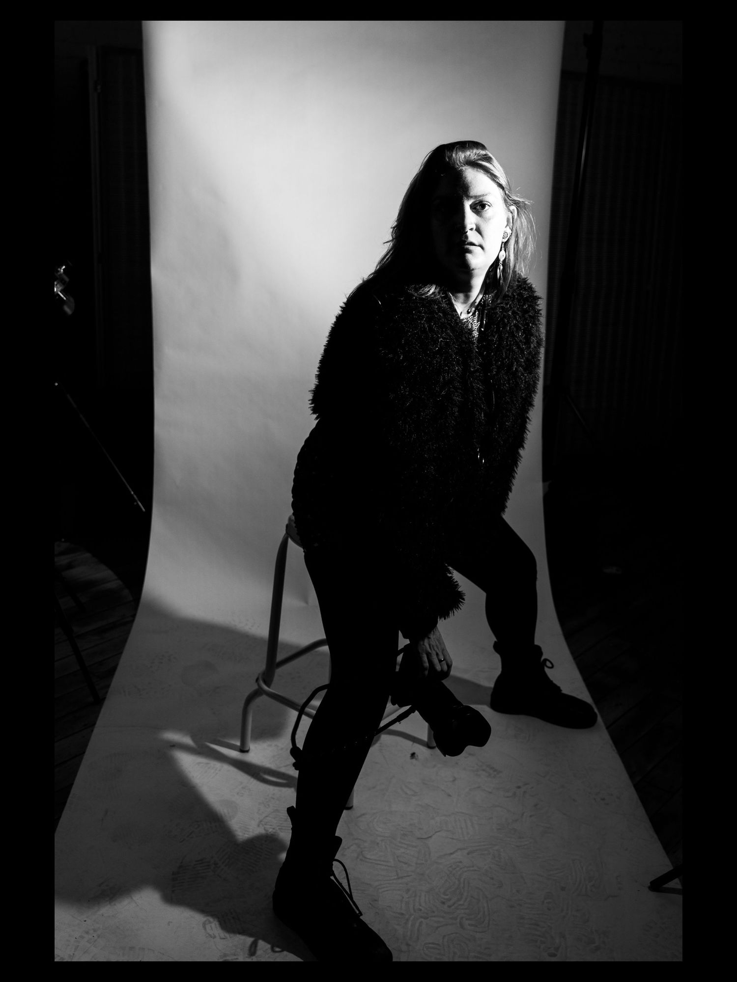 Black and white studio portrait of the artist in a fluffy black coat seated at a sharp angle on a stool, illuminated by dramatic lighting.