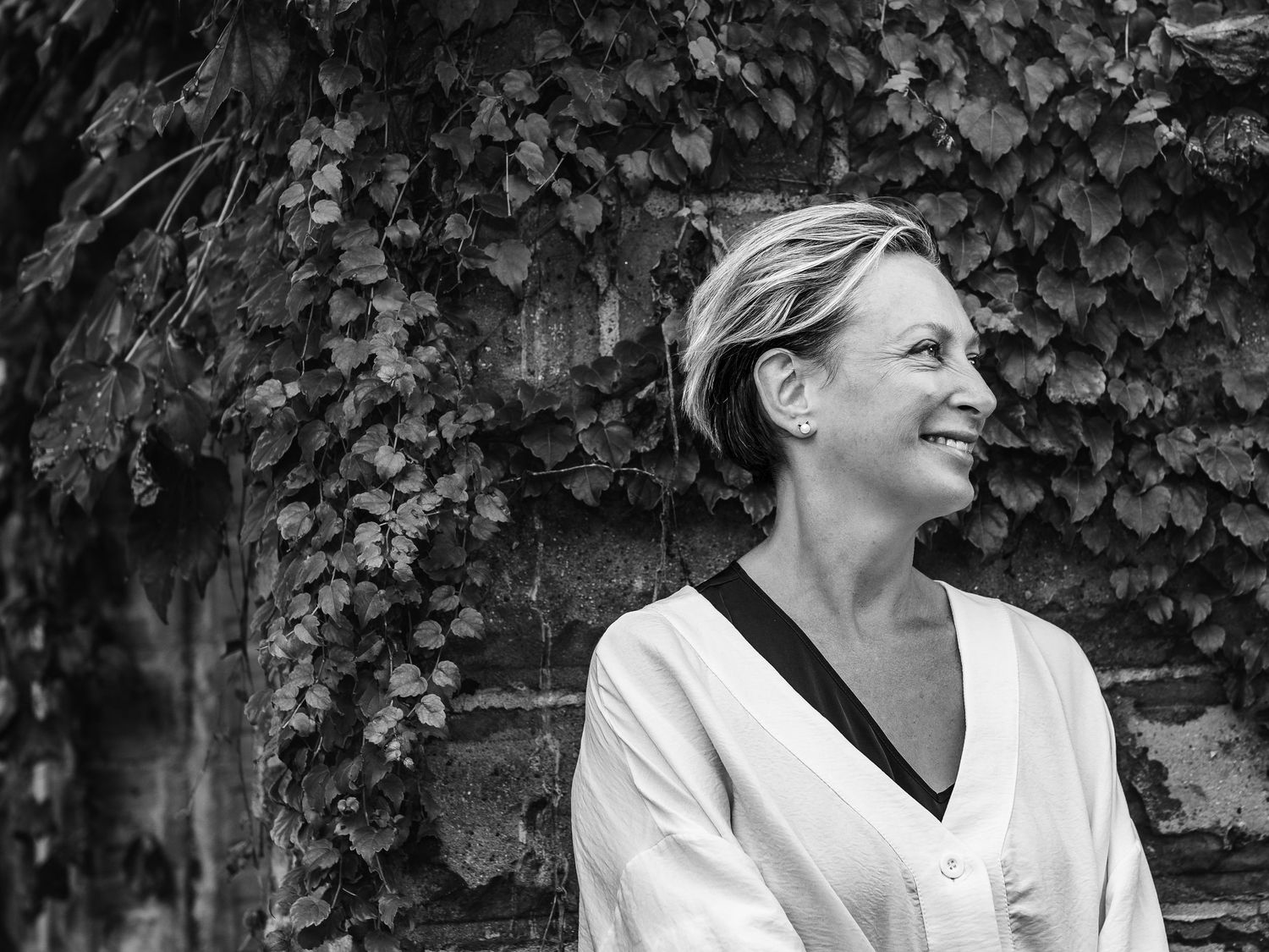 Black and white portrait of a smiling blonde woman standing against an ivy-covered stone wall outdoors.
