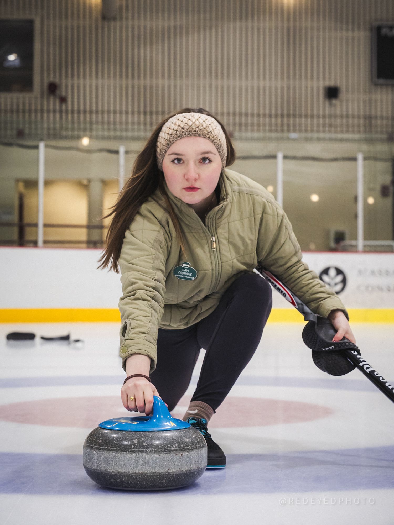 A curler delivers a stone down the ice while wearing an olive green jacket and black pants during a curling match.