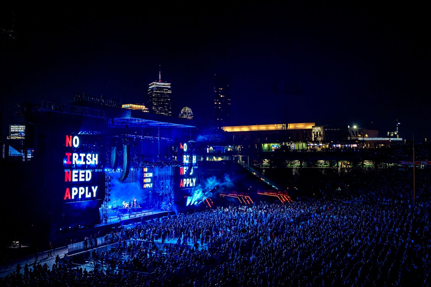 Large concert crowd at outdoor venue illuminated by blue lights with illuminated text display on building at night.