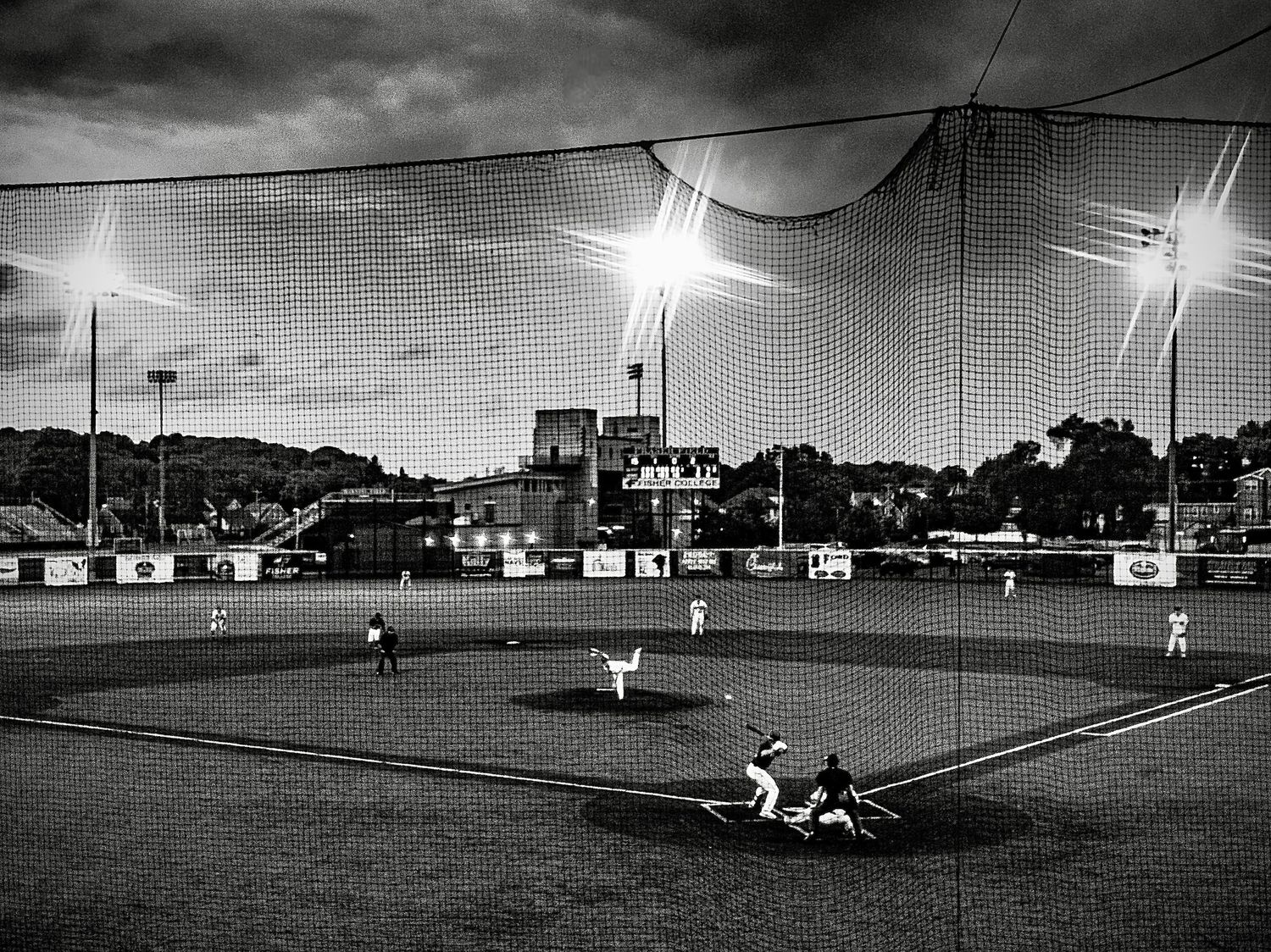 Black and white photo of a nighttime baseball game showing field, netting, stadium lights and players in action. Boston event photography.