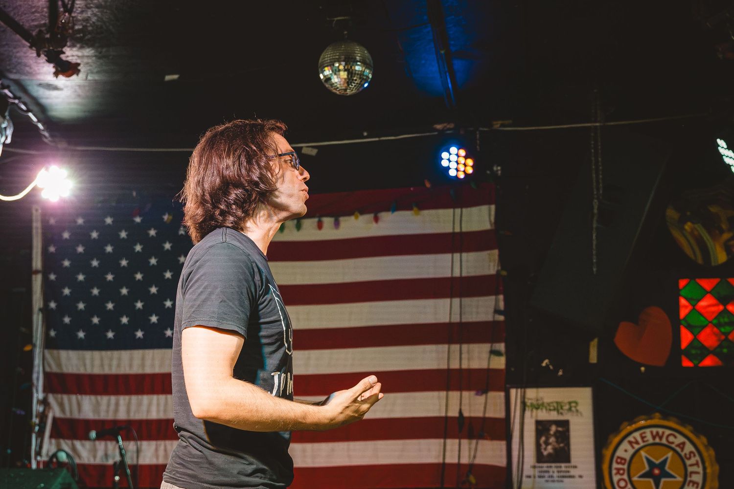 Man speaking on stage before an American flag with colorful stage lighting in a dark venue.