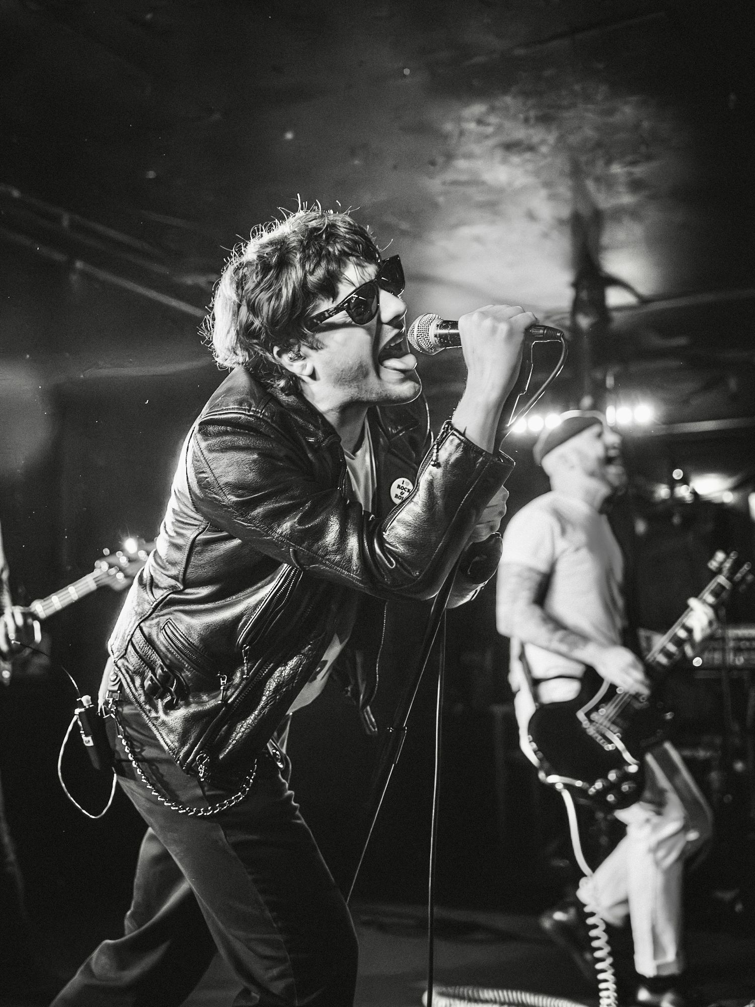 Black and white photo of a rock singer in leather jacket and sunglasses performing on stage with guitarist behind. (Band: Hell Beach)