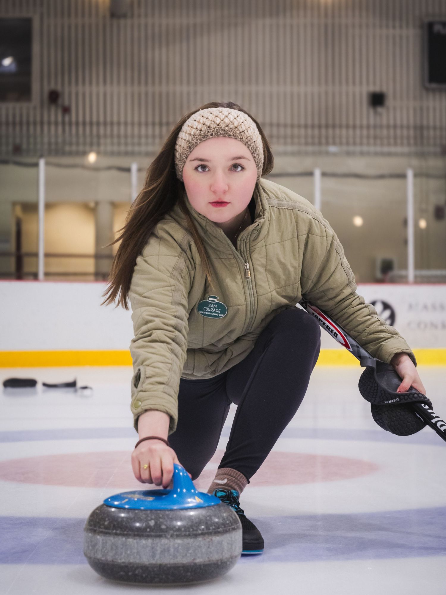 Young woman in green jacket and knit headband releases a blue curling stone on an indoor ice rink.