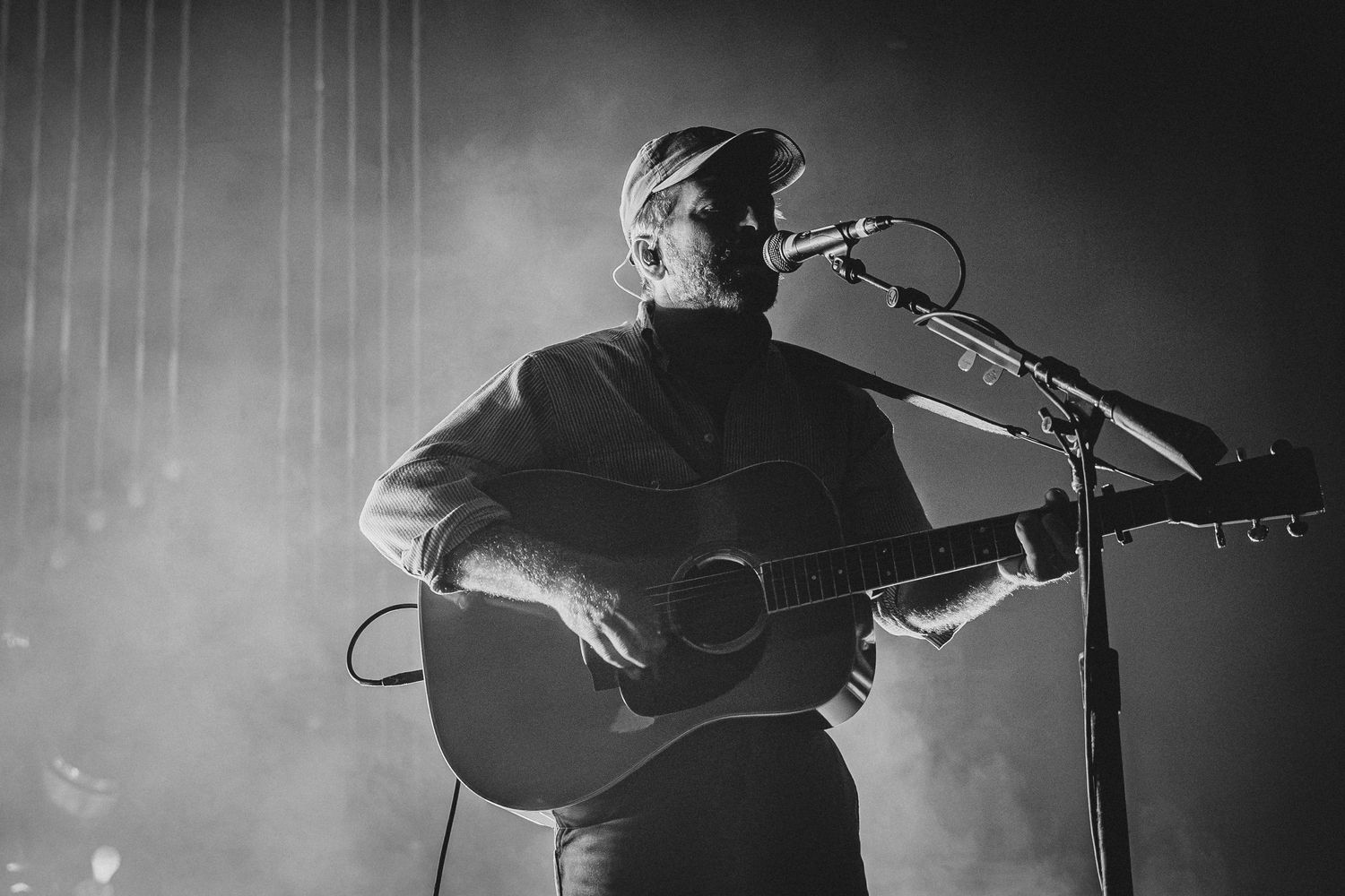 Black and white photo of a male guitarist performing on stage with acoustic guitar and microphone in dramatic lighting.