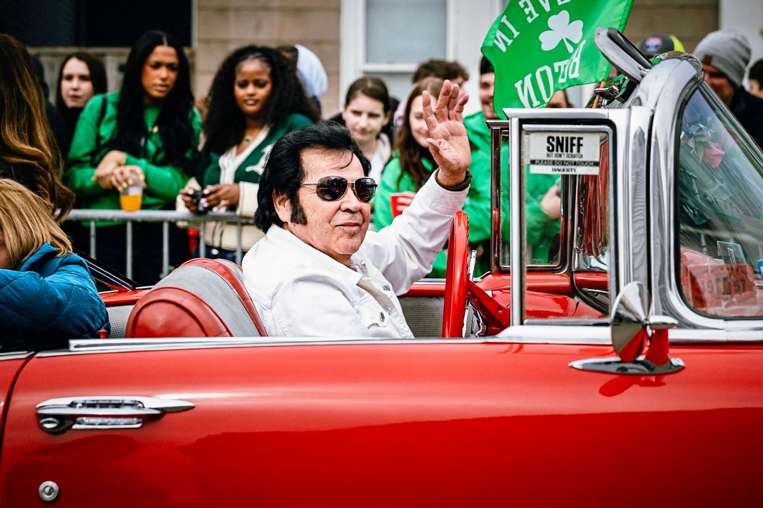 A classic red convertible car drives in a parade as spectators watch from behind barriers with green decorations visible.