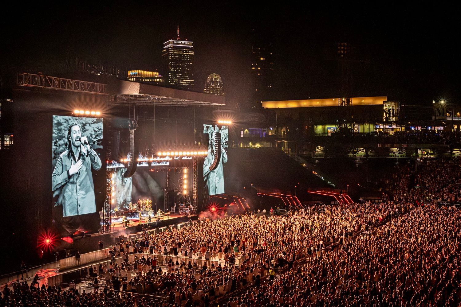 Large concert arena at night with illuminated video screens and massive crowd gathered for outdoor performance.