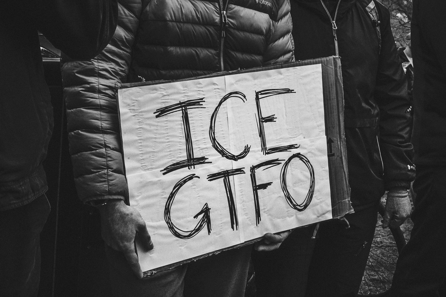 A protest sign with the text 'ICE GTFO' held by someone wearing a black winter coat.