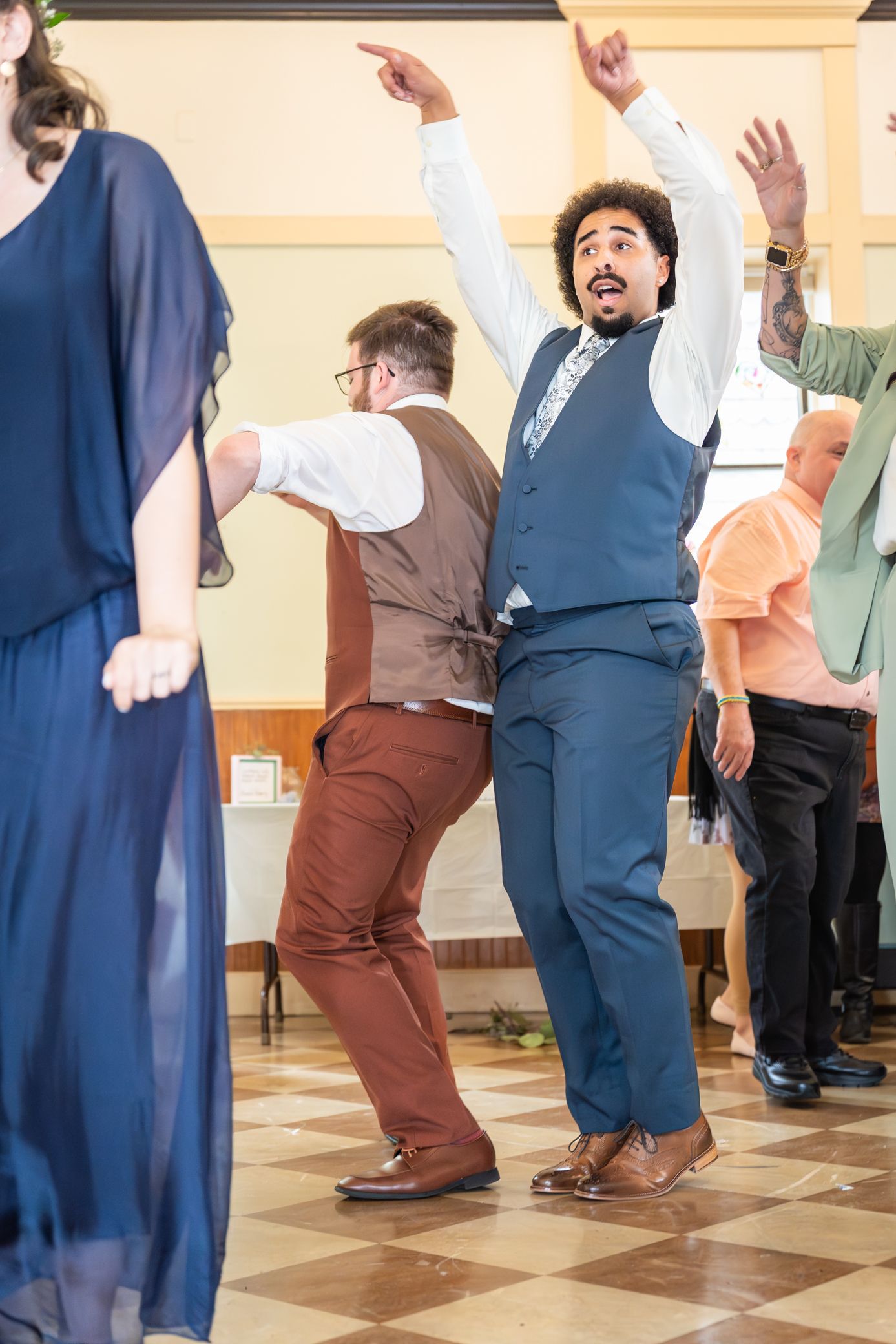 A group of people dancing in a line with their arms raised, wearing casual and formal attire on a tiled floor.