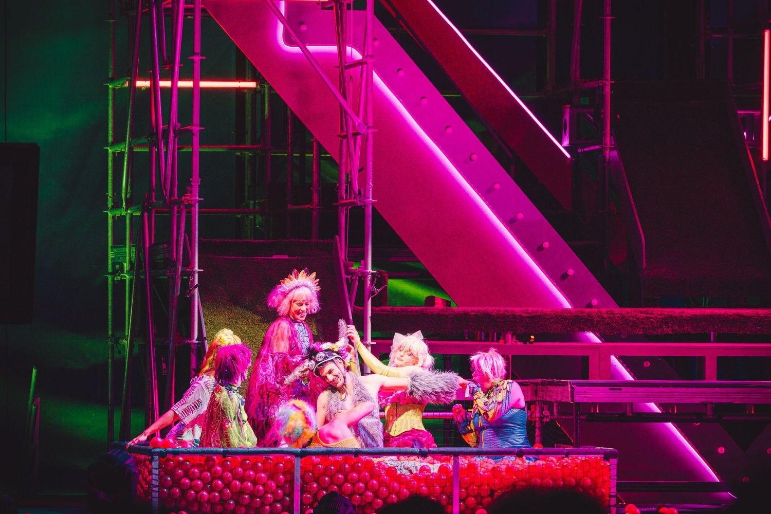 Colorful theatrical performers on stage bathed in pink neon lights during a vibrant musical production.