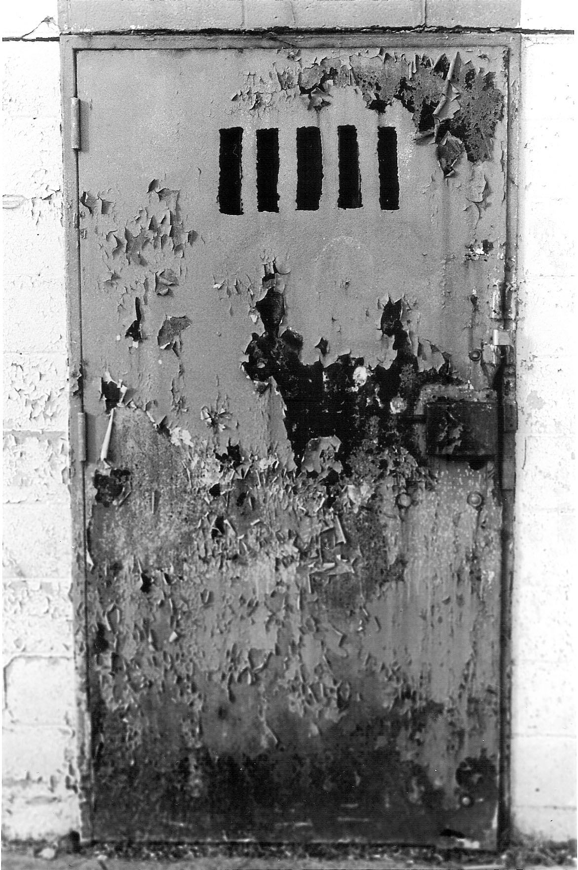 An old weathered metal prison cell door with five ventilation slits shows peeling paint and deterioration.