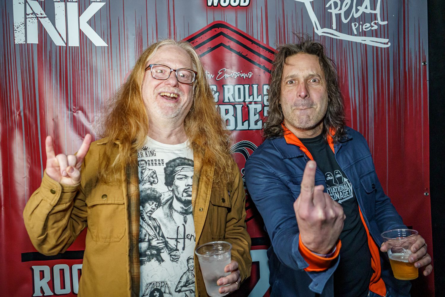 Two music fans pose together at a Rock and Roll event, with one showing a peace sign and another making a rock gesture.