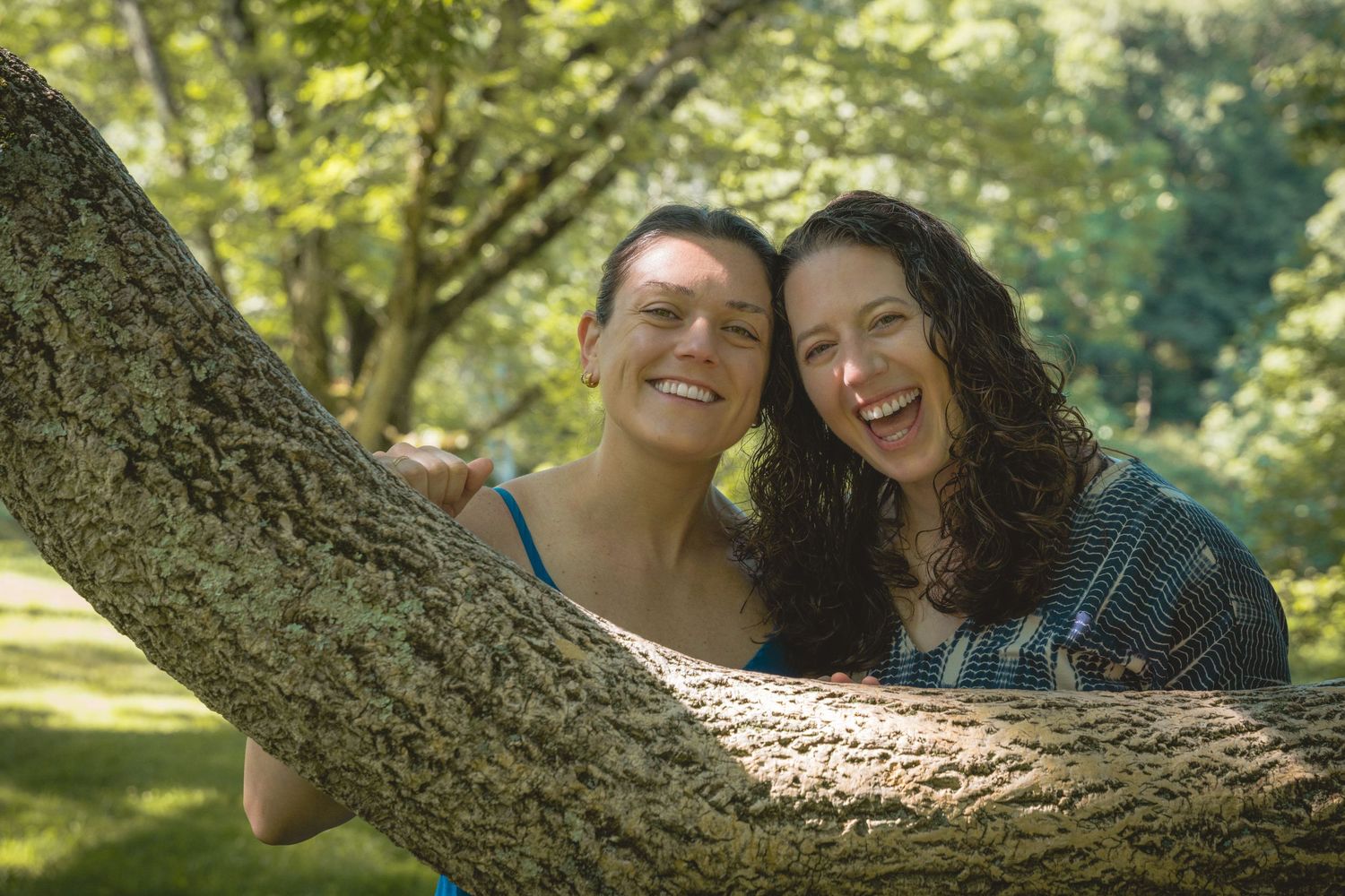 Two smiling women pose together behind a tree trunk in a lush green park on a sunny day.