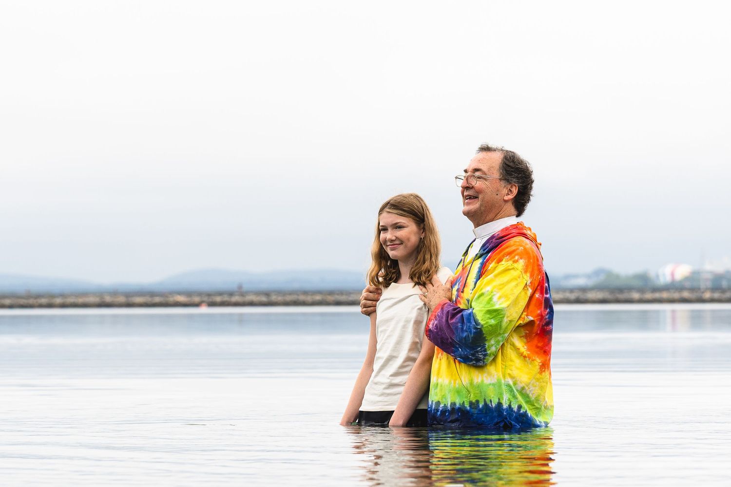 Two people laughing and standing waist-deep in calm water, one wearing a colorful tie-dye shirt.