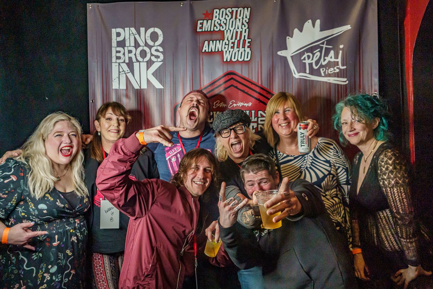 A group poses together showing excitement at a Pino Bros Ink event with signage and dark ambiance in the background.