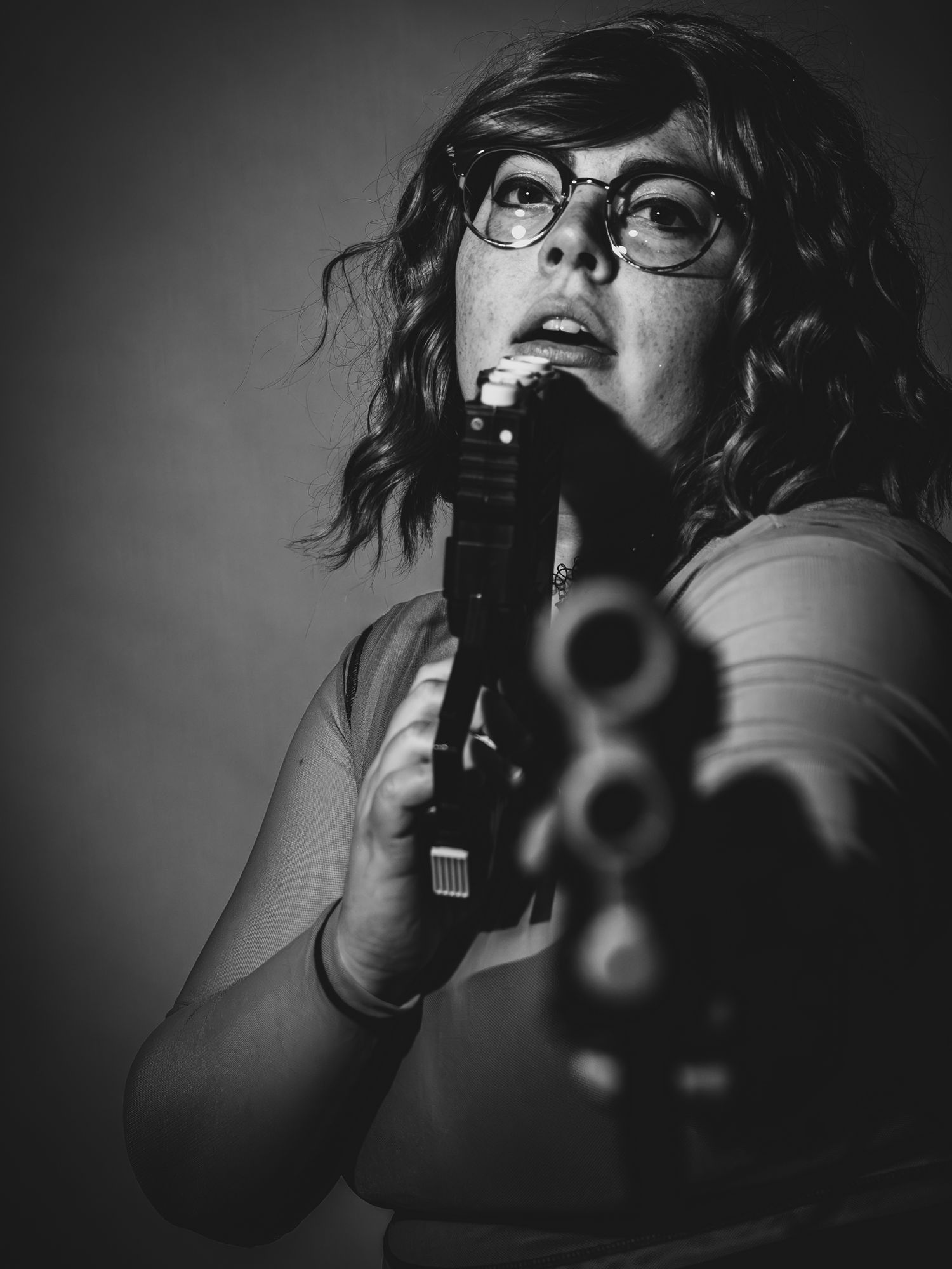Black and white portrait of a woman with glasses aiming a revolver toward camera in dramatic lighting.