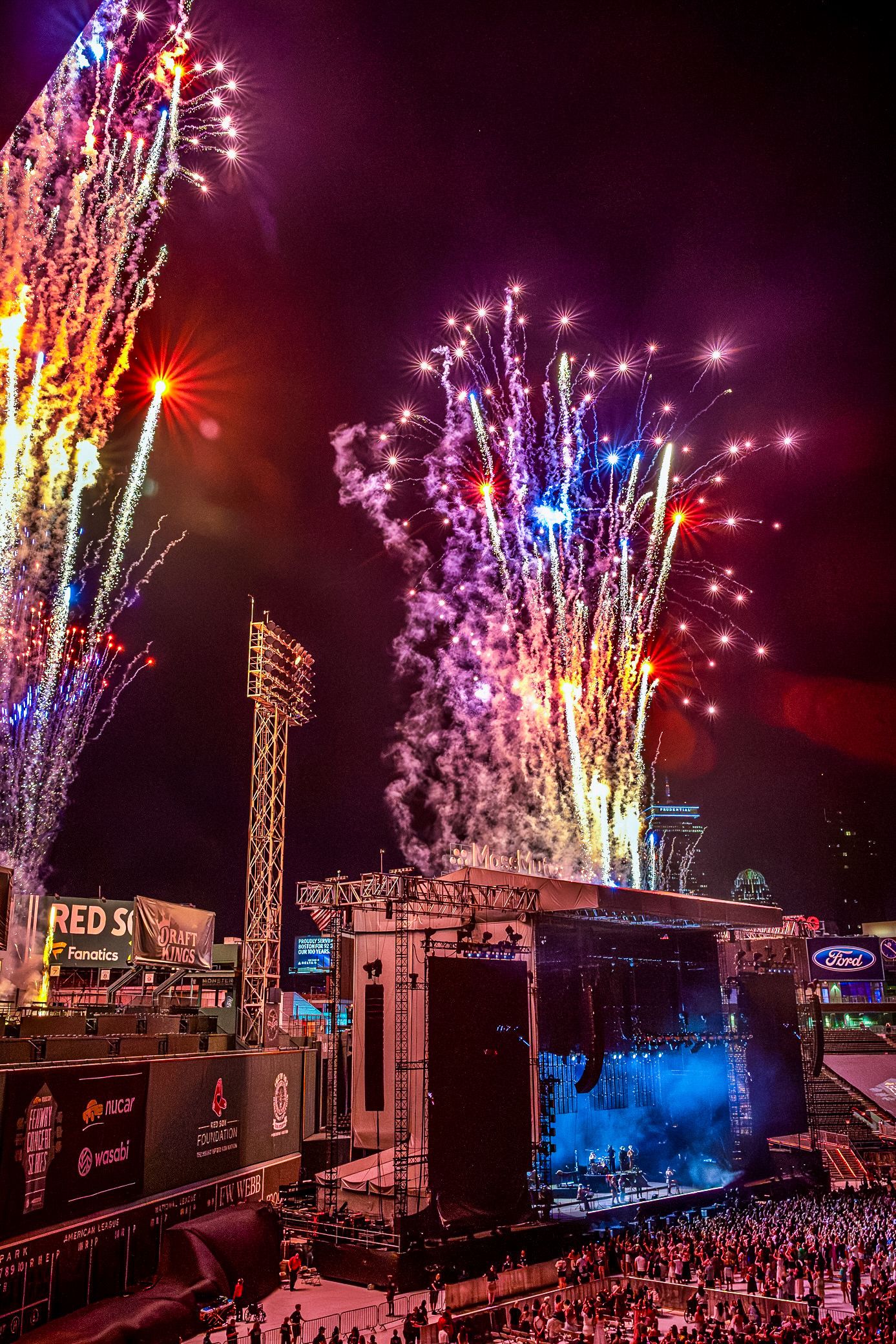 Spectacular fireworks display illuminates the night sky above an outdoor concert venue with multiple stadium lights.