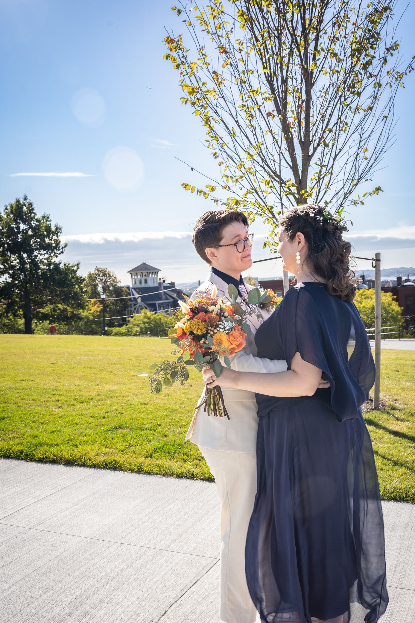 A couple embraces outdoors on their wedding day with autumn trees and blue sky in the background.